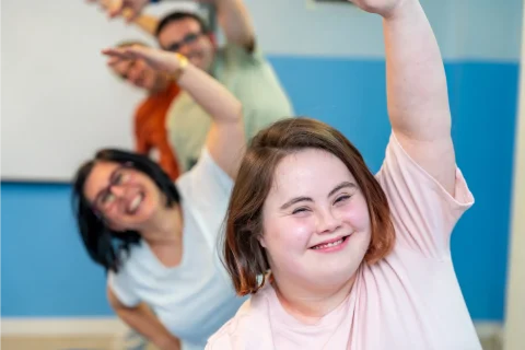 A young girls in front of a line of people doing a dance with their left arms in the air.
