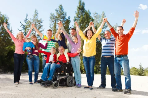A row of people holding hands, arms raised in the air. A man, wearing a red sweatshirt, in a mobility chair sits in front of the group.