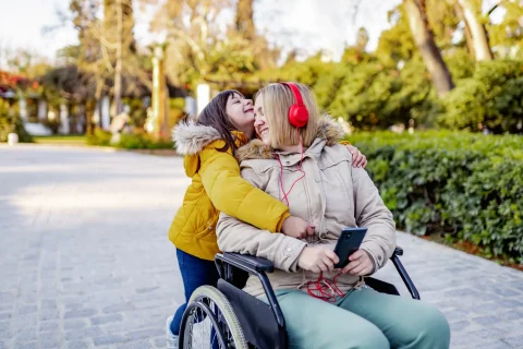 A woman in a wheelchair wearing red headphones is hugged by a young girl who's wearing a bright yellow jacket