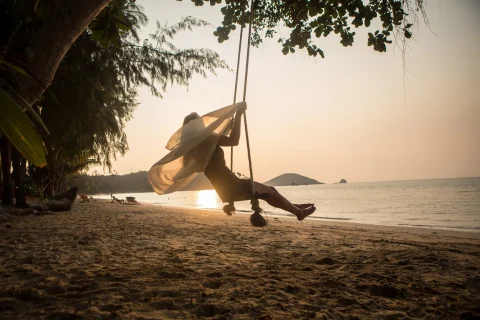 A woman with a white shawl swings above a sandy beach near the water's edge