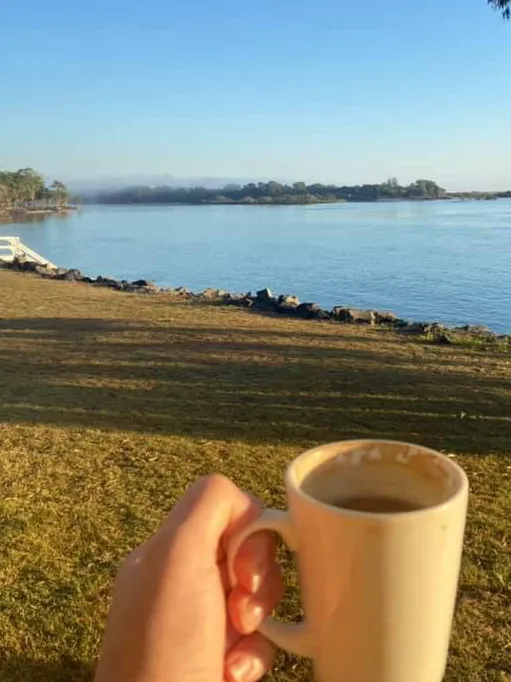 A hand holding a mug of coffee outside with a view of the foreshore.