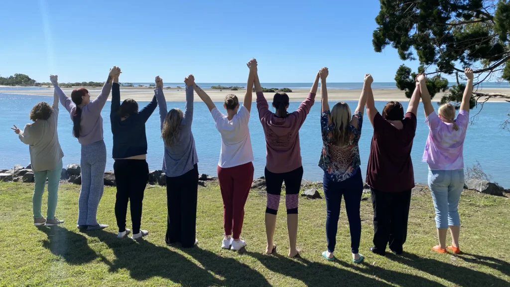 A group of people with their backs to the camera, they're standing in a line with held hands raised in the air, all looking across an inlet towards the open ocean.